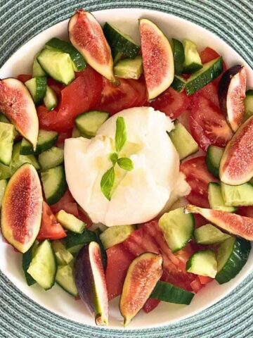 A white serving bowl containing sliced purple figs, crisp green cucumbers, ripe tomatoes, and creamy burrata cheese.